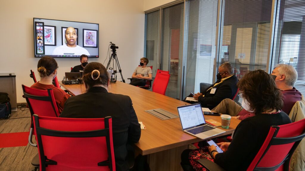 Several people sit around a conference table watching a virtual presentation on a screen mounted on the wall.