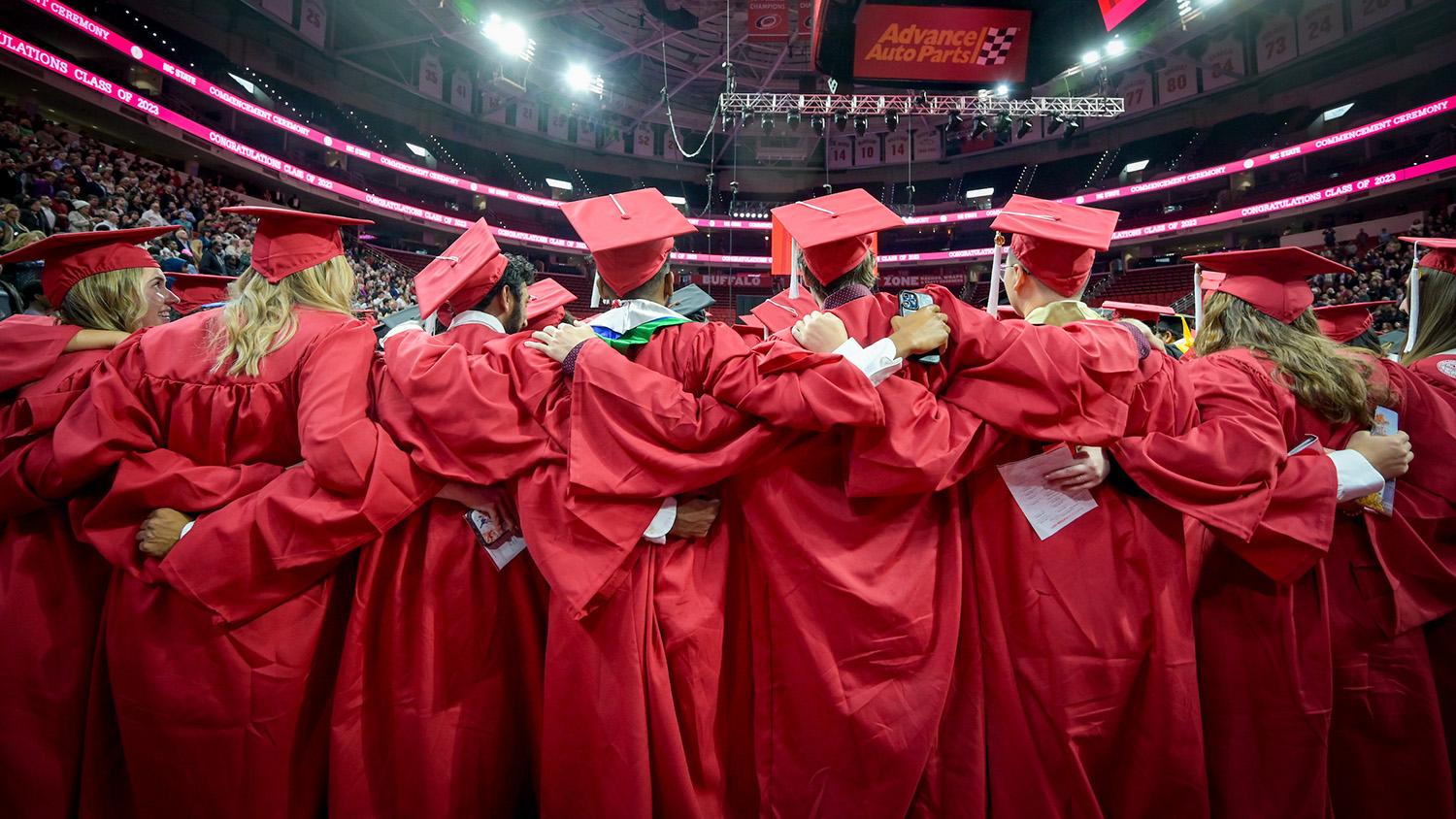 fall NC State graduates with their arms around one another while singing the alma mater