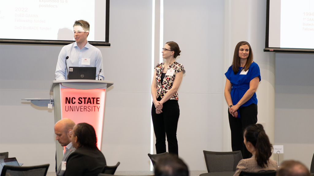 Three faculty members from the BIT Program stand on stage addressing the audience.