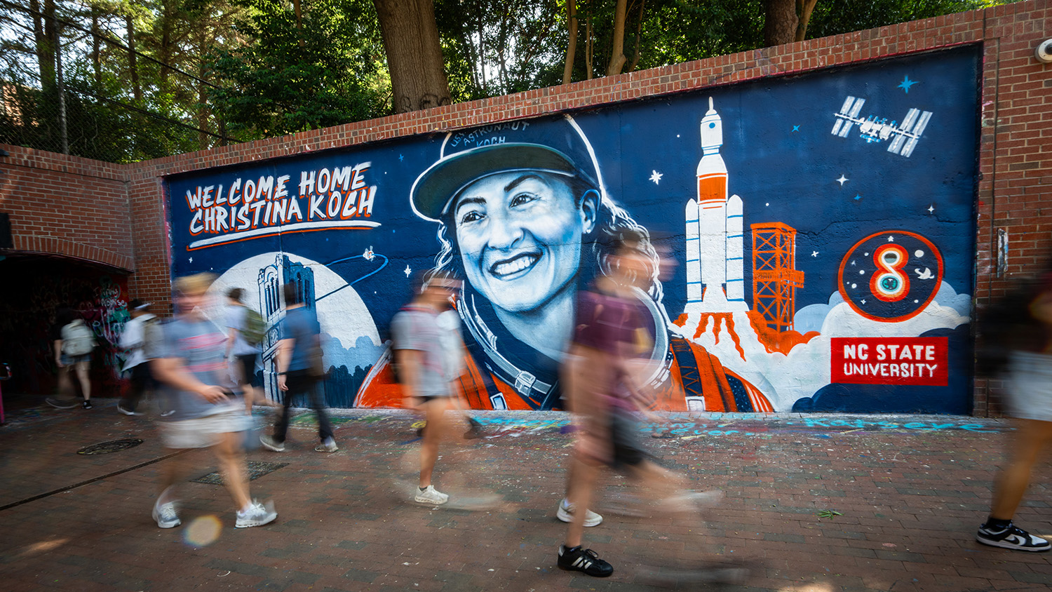 A vibrant mural welcomes Christina Koch home, depicting her as an astronaut with rockets and the International Space Station, set against a brick wall as people walk by.
