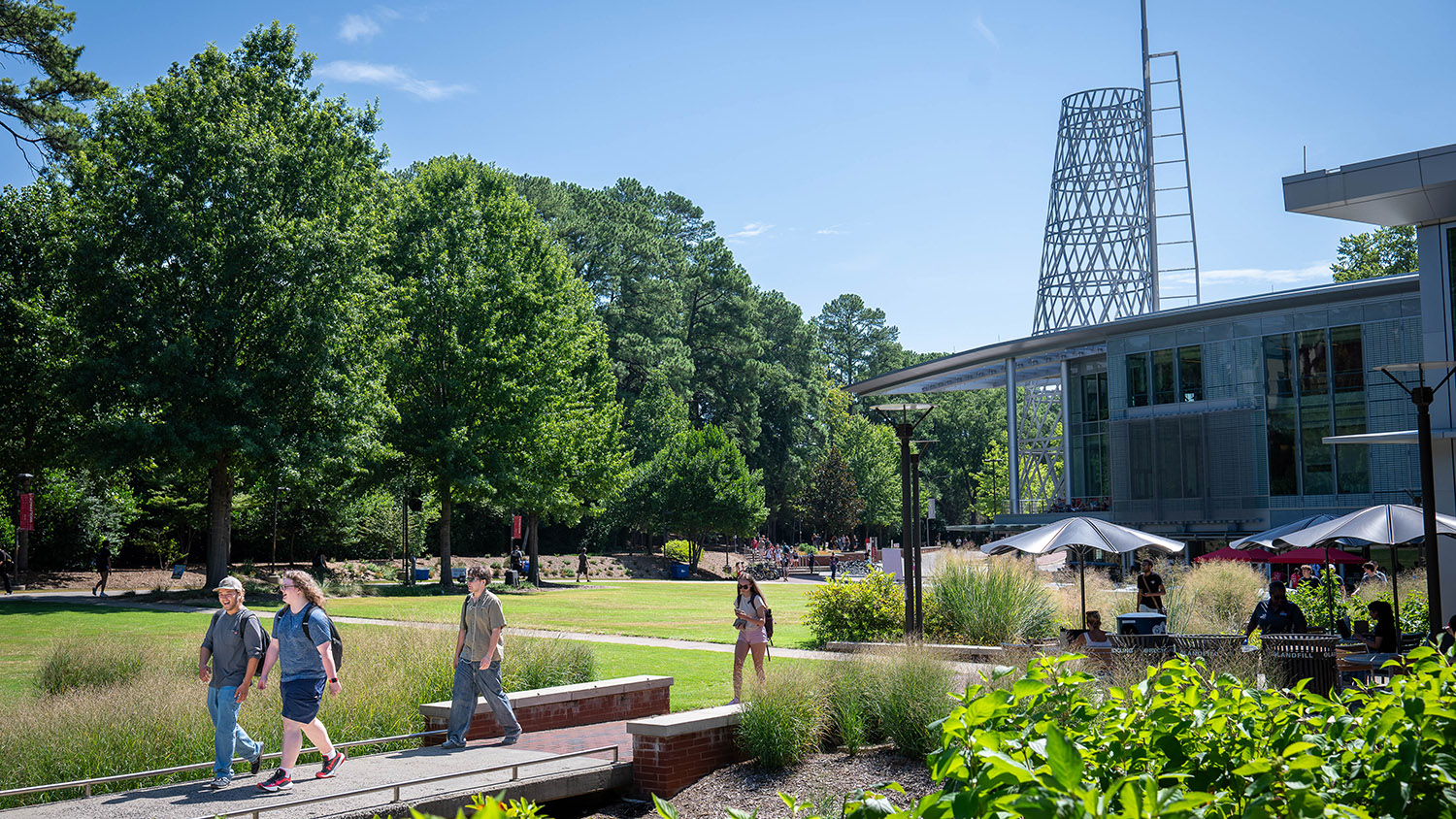 People stroll across a wooden bridge in a sunlit campus garden, with modern architecture and a distinctive tower visible in the background.