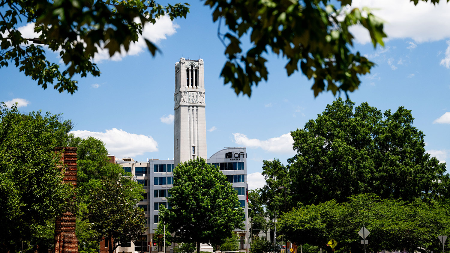 A historic clock tower stands tall behind modern buildings, framed by lush green trees under a bright blue sky.